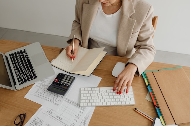 Woman at a desk with papers, computer and calculator working in a notebook